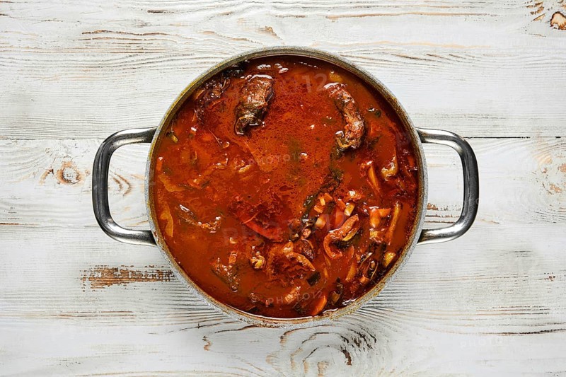 Overhead view of a silver pot with two handles on a white wooden table. The pot is filled with a rich beef stew, filled with chunks of meat and vegetables.