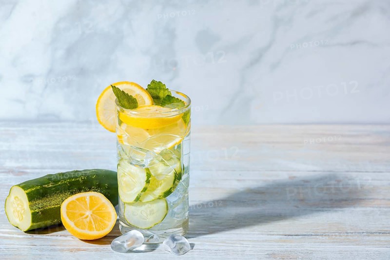 A glass of cucumber water with lemon slices, mint leaves and ice cube cubes, cucumber and lemon on a table next to it