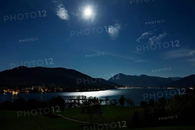 Night scene with moonlight over a calm lake, surrounded by mountain silhouettes and shining lights on the shore, Tegernsee