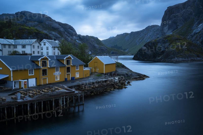 View of Nusfjord with yellow or ochre-coloured and white houses by the sea, mountains in the background. At night at the time of the midnight sun. Cloudy sky. Long exposure with blurred clouds. Early summer. Nusfjord, Flakstadoya, Lofoten, Norway, Europe