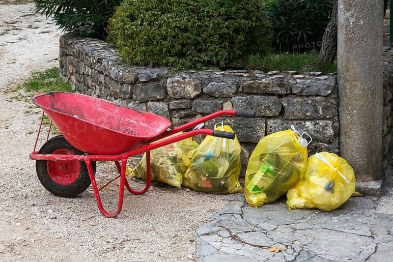 Red metal wheelbarrow next to translucent yellow plastic thrash bags filled with various discarded domestic garbage, Salona, Solin, Croatia, Europe