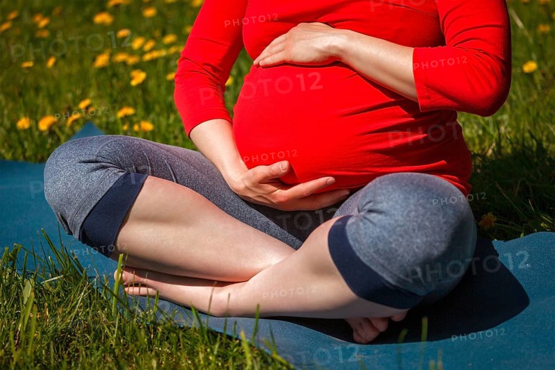 Pregnancy yoga exercise, pregnant woman doing asana Sukhasana easy yoga pose holding her abdomen outdoors on grass lawn with dandelions in summer