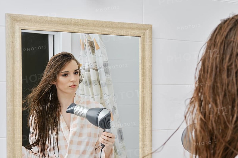 Young woman wearing bathrobe blow drying long hair after shower and looking in mirror, good-looking female with hair dryer in bathroom. Haircare and beauty concept