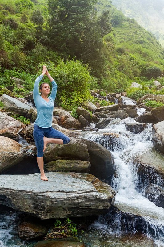 Woman in Hatha yoga balance yoga asana Vrikshasana tree pose at waterfall outdoors