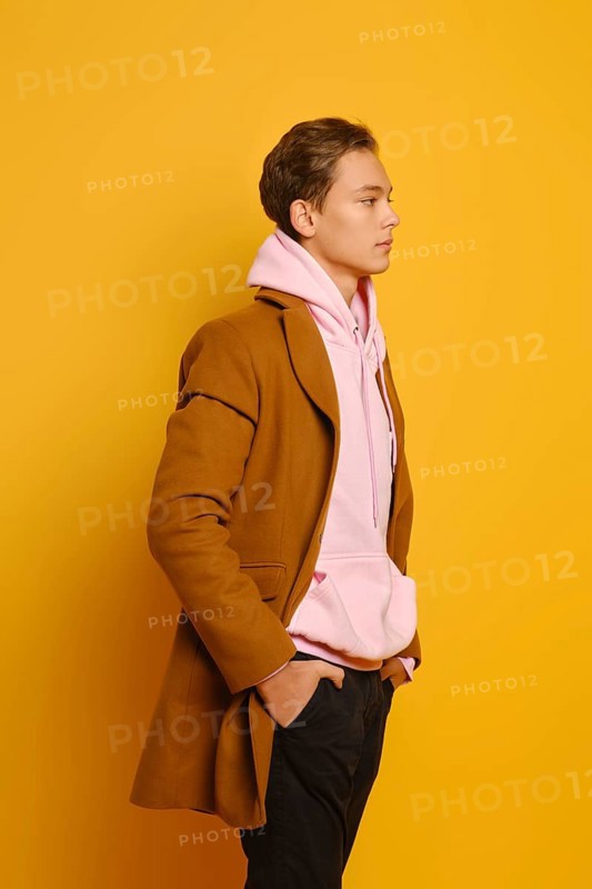 Handsome stylish man in brown trenchcoat and pink sweatshirt with hood standing in profile over yellow studio background
