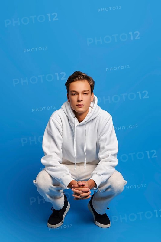 Attractive man wearing white sweatshirt and sweatpants is squatting over blue studio background