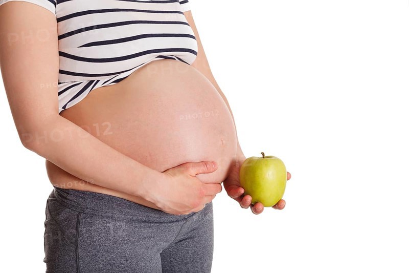 Healthy eating and diet during pregnancy concept, pregnant woman standing and holding green apple near her stomach isolated on white