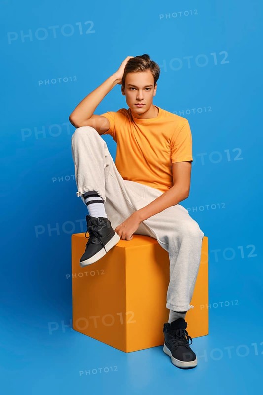 Positive man in orange undershirt and white sweatpants sits on wooden cube over blue studio background