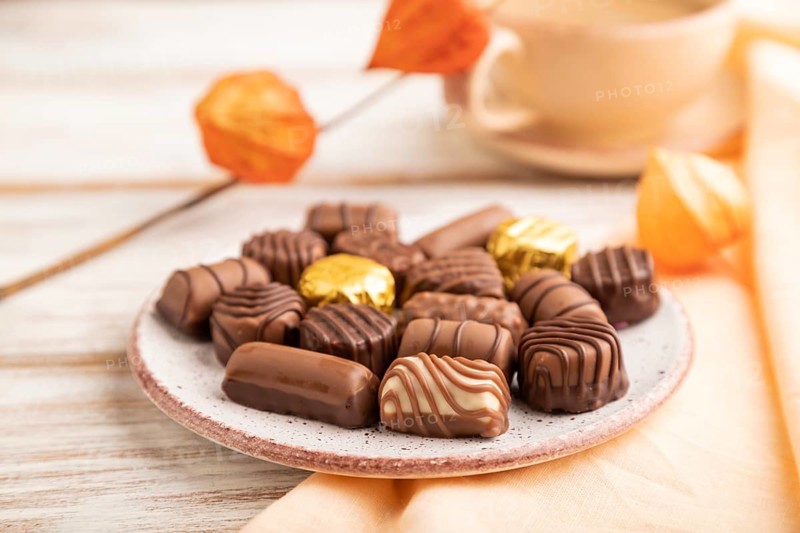 Chocolate candies with cup of coffee and physalis flowers on a white wooden background and orange textile. side view, close up, selective focus