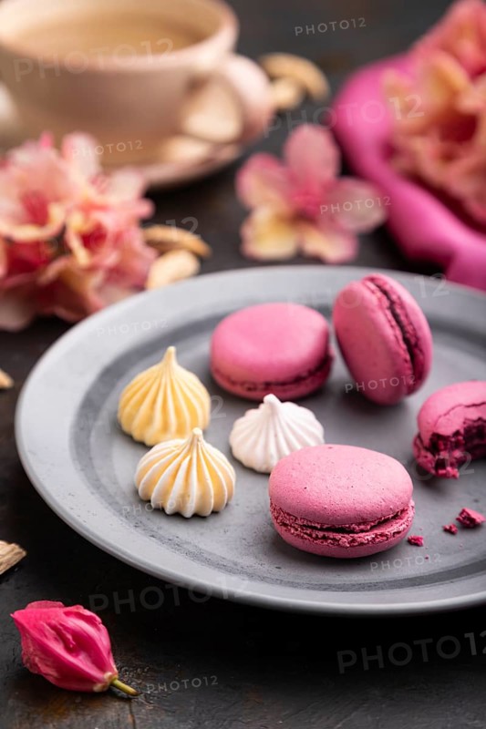 Purple macarons or macaroons cakes with cup of coffee on a black concrete background and pink textile. Side view, close up, selective focus