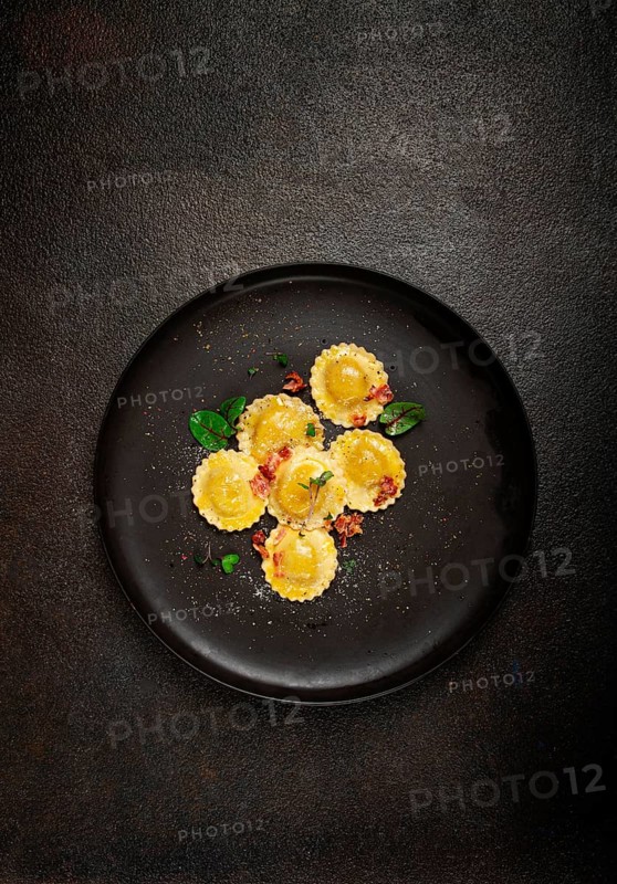 Fresh ravioli, with butter, spices and herbs, fried bacon, on a black plate, top view, dark background, no people