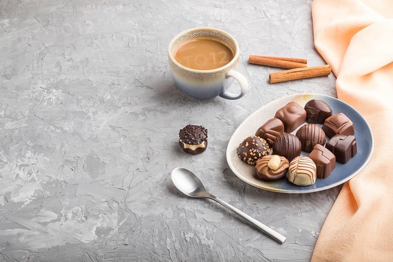 Different chocolate candies and a cup of coffee on a gray concrete background and orange textile. side view, copy space