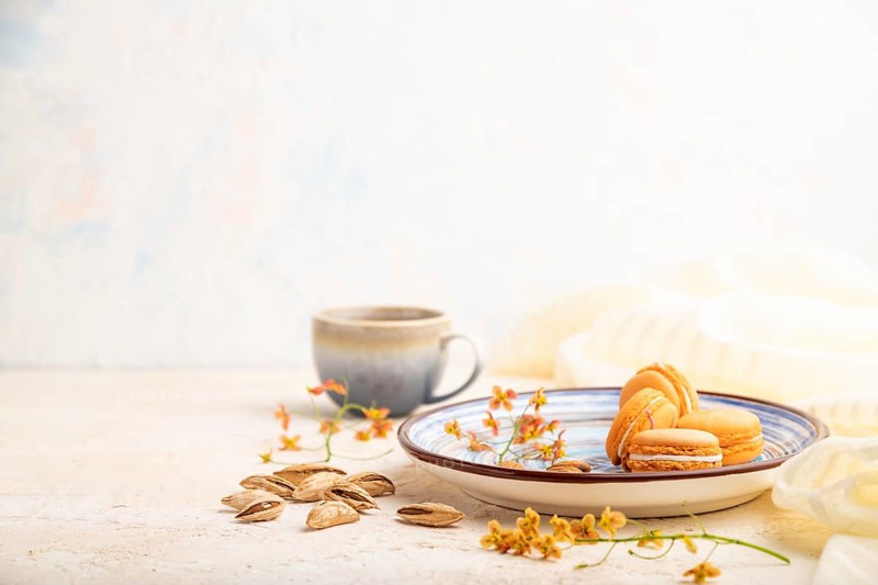 Orange macarons or macaroons cakes with cup of coffee on a white concrete background and linen textile. Side view, copy space