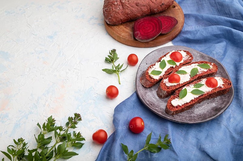 Red beet bread sandwiches with cream cheese and tomatoes on white concrete background and blue linen textile. Side view, copy space, close up