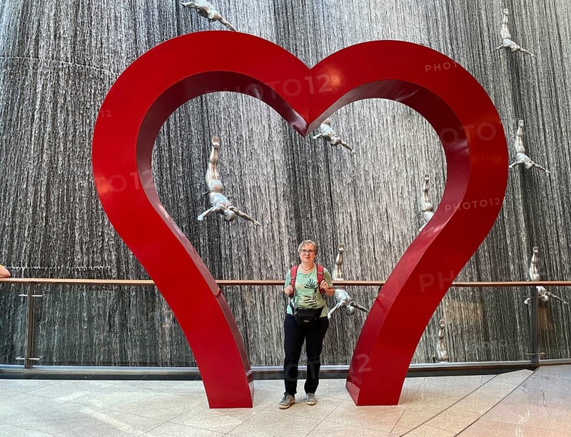 Interior shot, best ager, elderly woman, standing in frame, heart, in front of The Waterfall, artificial indoor waterfall with life-size jumper sculptures, installation on the pearl diving tradition, Dubai Luxury Shopping Mall, luxury shopping centre, Downtown, Dubai, United Arab Emirates, VAR, Asia
