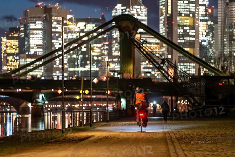 Skyline of the city centre of Frankfurt am Main, cyclist with light on the cycle path, pavement, promenade along the river Main, dusk, Hesse, Germany raft bridge in the background