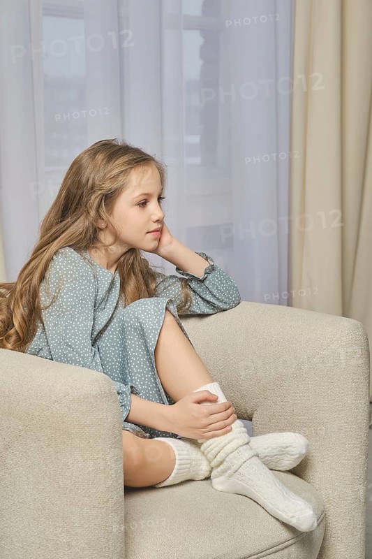 A young pensive girl sits in profile on a soft armchair in a well-lit room with her hand resting her chin. She is wearing a pale green dress and warm knitted socks