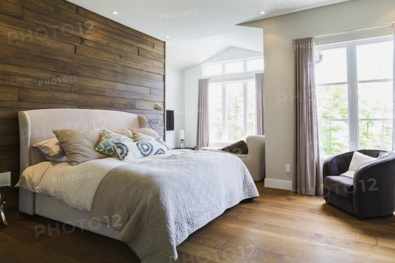 King size bed with grey linen cloth headboard, charcoal velour armchair in master bedroom with engineered hickory wood floorboards inside a luxurious contemporary bungalow style home, Quebec, Canada, North America