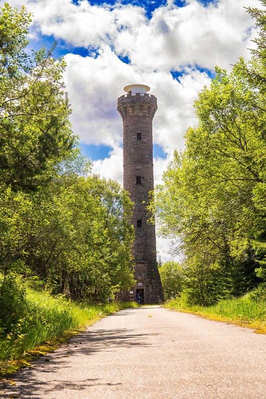 A tall stone tower rises above a road, flanked by trees under a sky full of clouds and sunbeams, Hohlohturm, Bad Wildbad, Black Forest, Germany, Europe