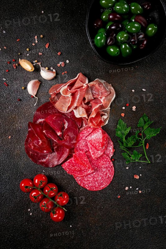Assorted dried meat, fresh tomatoes and olives spread out on a dark surface, food composition, top view, no people