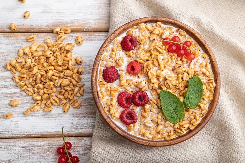 Wheat flakes porridge with milk, raspberry and currant in wooden bowl on white wooden background and linen textile. Top view, flat lay, close up
