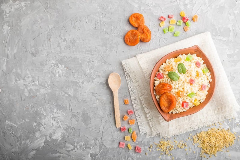 Bulgur porridge with dried apricots and candied fruits in clay bowl on a gray concrete background and linen textile. Top view, flat lay, copy space. Turkish traditional cuisine