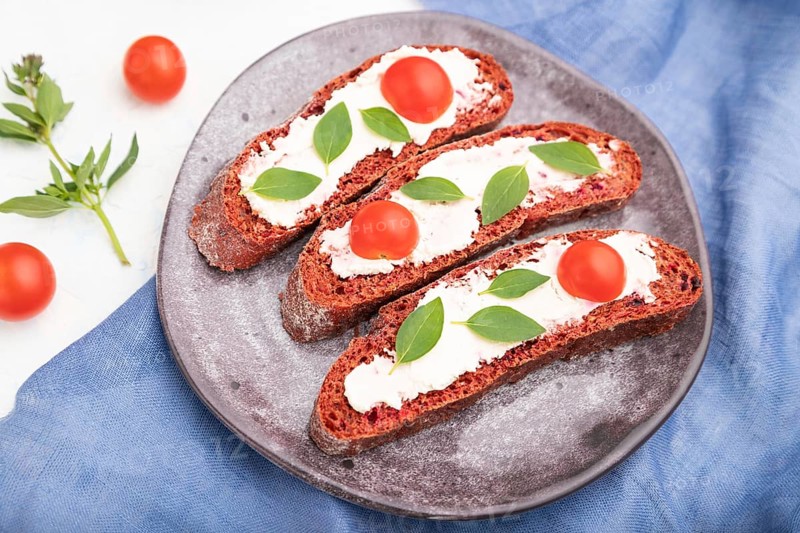 Red beet bread sandwiches with cream cheese and tomatoes on white concrete background and blue linen textile. Side view, selective focus, close up