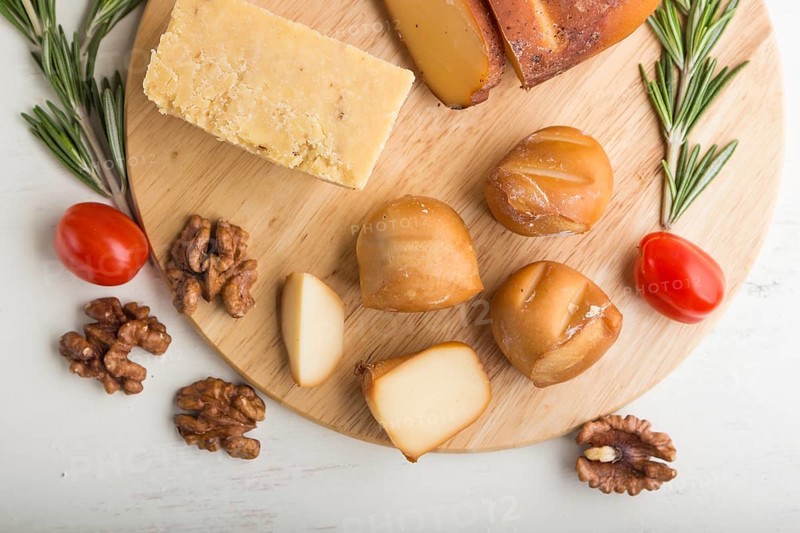 Smoked cheese and various types of cheese with rosemary and tomatoes on wooden board on a white wooden background. Top view, flat lay, close up