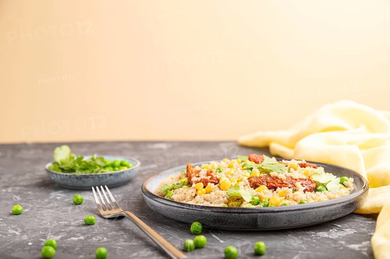 Quinoa porridge with green pea, corn and dried tomatoes on ceramic plate on a gray and orange background and yellow textile. Side view, copy space