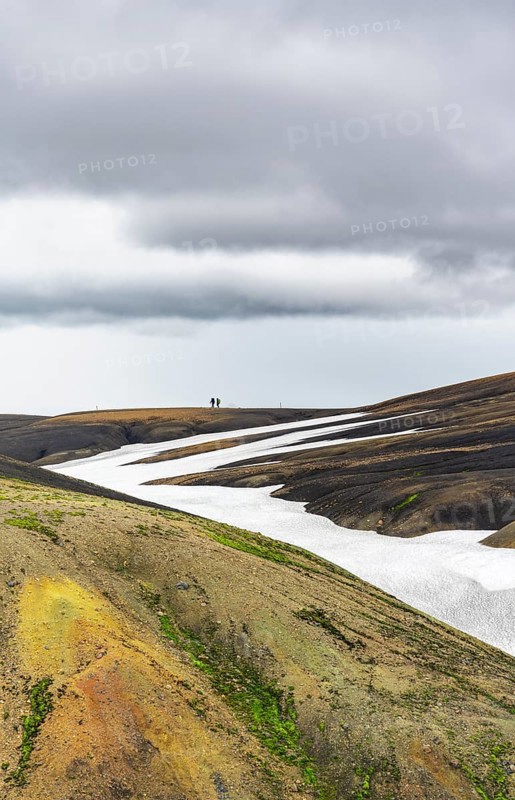 Two hikers on a hill in the distance, colourful volcanic landscape with hills and snow, Laugavegur trekking trail, Landmannalaugar, Fjallabak Nature Reserve, Icelandic Highlands, Suðurland, Iceland, Europe