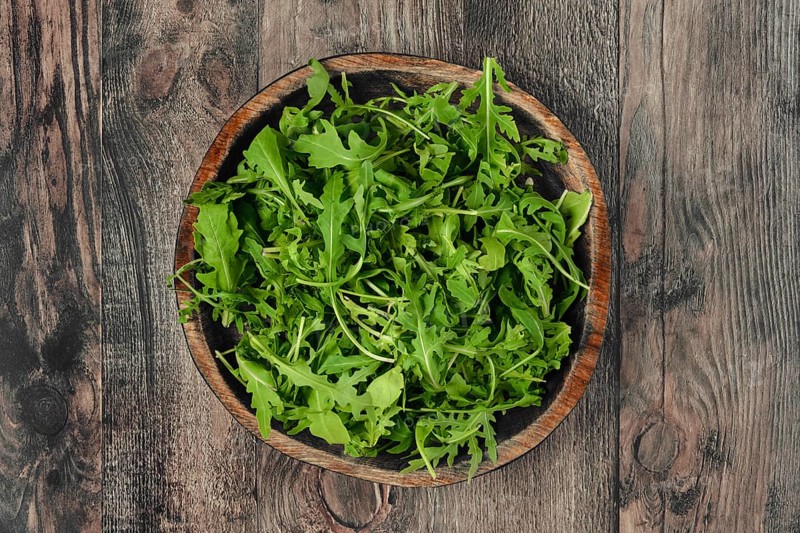 A rustic wooden bowl with fresh arugula leaves on a textured wooden table, showcasing vibrant greenery perfect for salads or garnishes. The natural setting emphasizes freshness