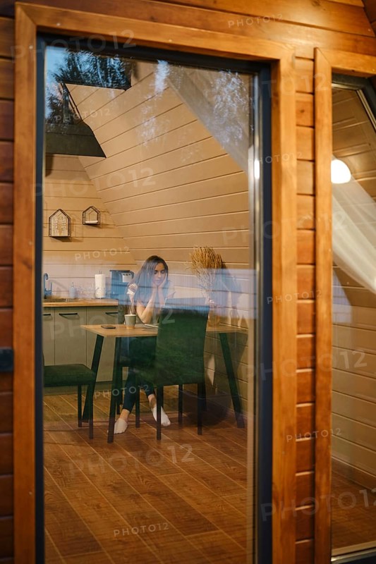 View through the large window at a pensive woman sitting at a table in tiny house in the evening