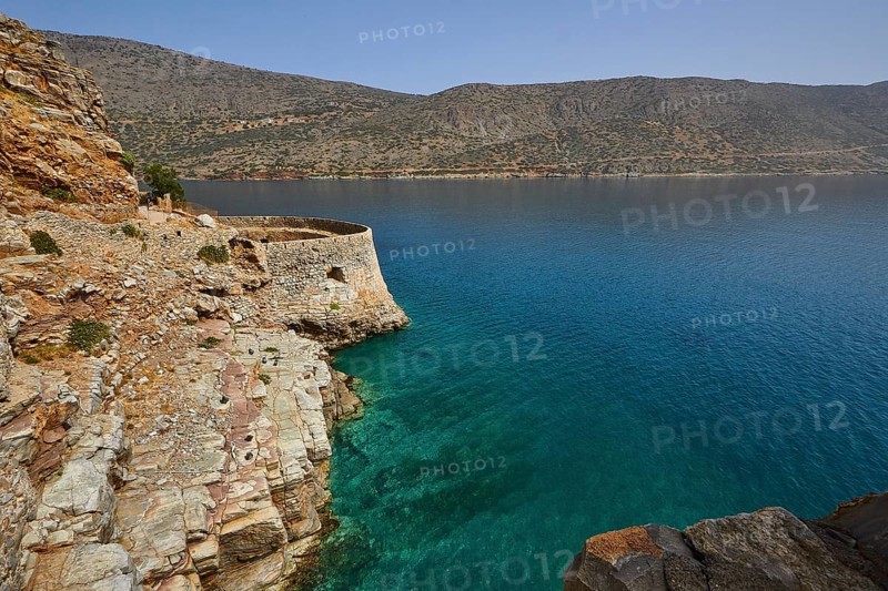 Rustic stone wall on cliffs above a turquoise sea with mountains in the distance, Venetian sea fortress, Leprosy Island, Spinalonga, Elounda Bay, Mirabello Bay, East Crete, Crete, Greek Islands, Greece, Europe