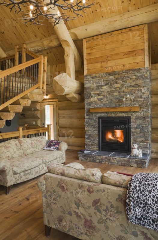 High angle view of twisting staircase with wooden and black wrought iron handrail and brown stained half log steps and lit natural stone fireplace in living room with hardwood floor and flowery motifs upholstered sofas inside handcrafted Eastern white pine Scandinavian log cabin home, Quebec, Canada, North America