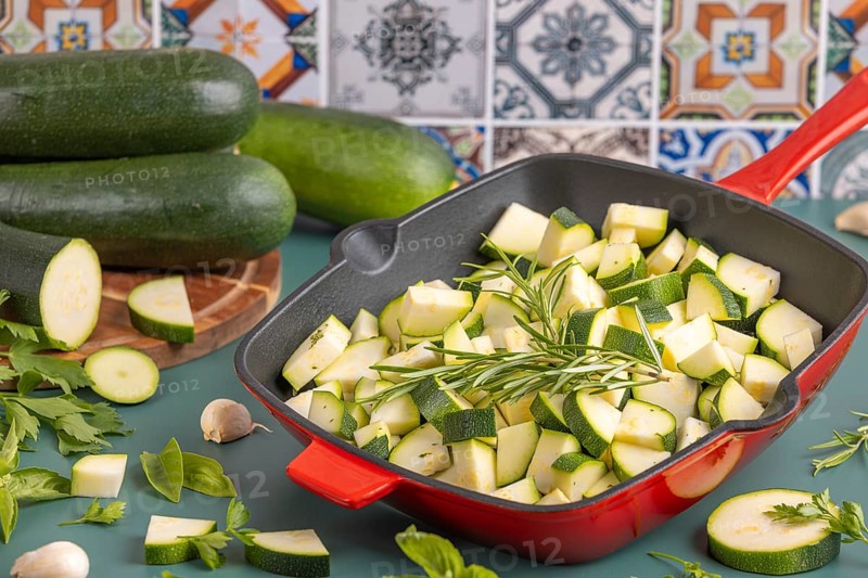 Diced courgettes in a red pan, surrounded by fresh herbs and whole courgettes in front of colourful tiles