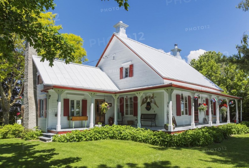 Old circa 1840 white horizontal wood plank with red trim Canadiana style house facade with silver standing seam sheet metal roof in early summer, Quebec, Canada, North America