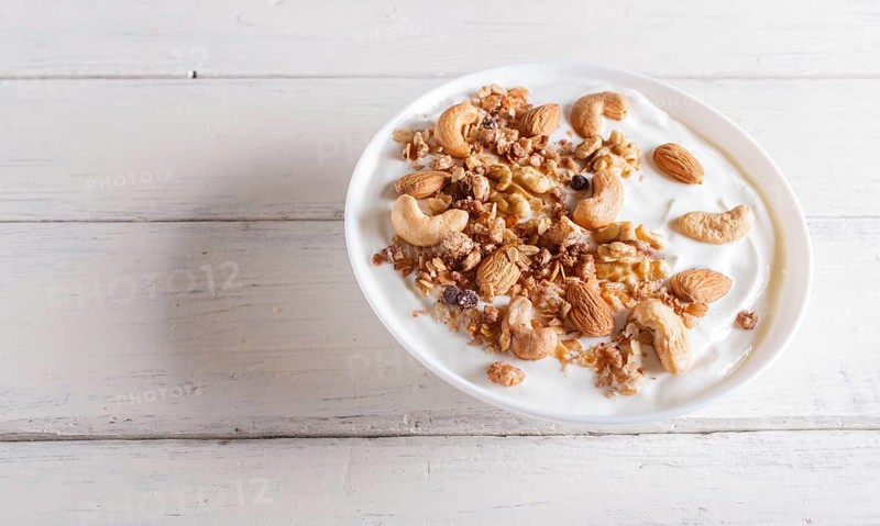 White plate with greek yogurt, granola, almond, cashew, walnuts on white wooden background. close up. copy space