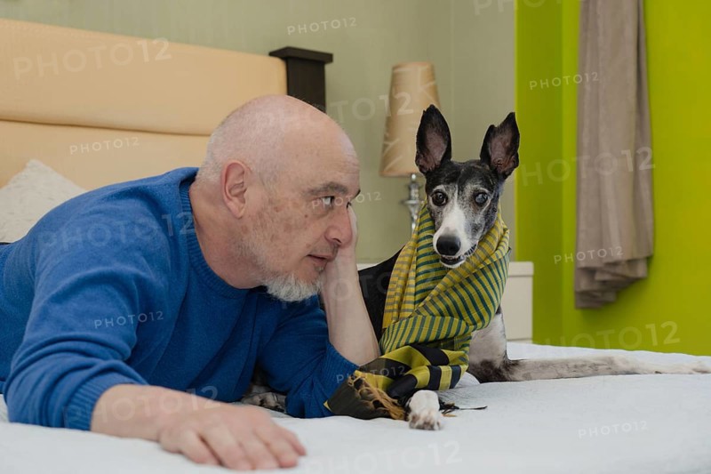 A mature gentleman, accompanied by his greyhound companion, poses for the camera from the comfort of his bed