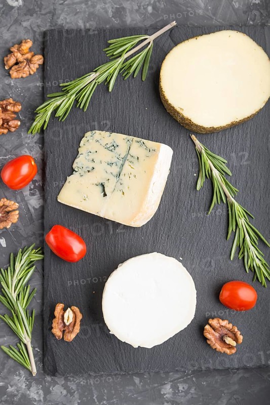 White goat cheese and various types of cheese with rosemary and tomatoes on black slate board on a black concrete background. Top view, close up, flat lay
