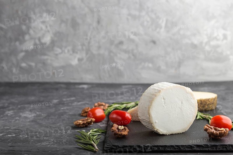 White goat cheese and various types of cheese with rosemary and tomatoes on black slate board on a black concrete background. Side view, copy space, selective focus