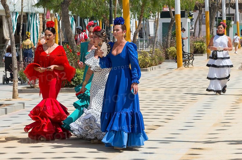 Five woman in different colourful flamenco dresses, with flowers in their hair, strolling on a festive street, Feria de la Manzanilla, Feria de Sanlúcar, Sanlúcar de Barrameda, Sanlucar, Cádiz, Cadiz, Andalusia, Spain, Europe