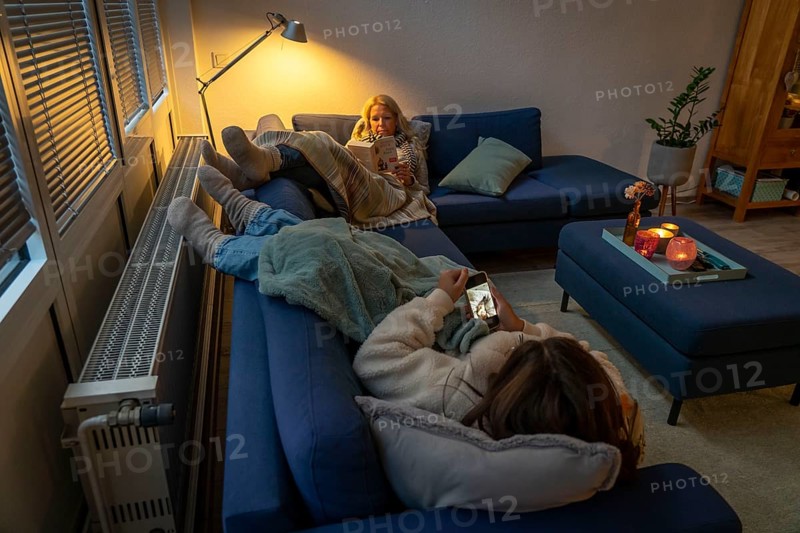 Symbolic image energy saving, cold winter, energy crisis, cold flat, mother and daughter lying on the sofa, warmly dressed, candlelight, heating on to a minimum, warming their feet on the radiator
