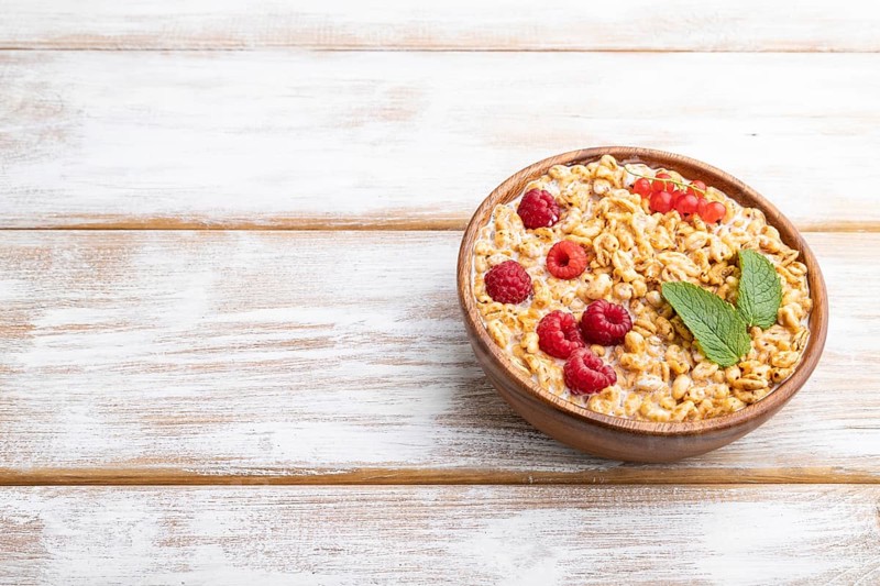 Wheat flakes porridge with milk, raspberry and currant in wooden bowl on white wooden background. Side view, copy space, close up