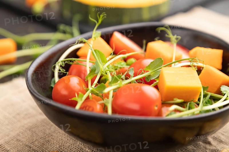 Vegetarian vegetable salad of tomatoes, pumpkin, microgreen pea sprouts on black concrete background and linen textile. Side view, close up, selective focus