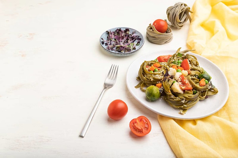 Tagliatelle green spinach pasta with tomato, pea and microgreen sprouts on a white wooden background and yellow textile. Side view, copy space