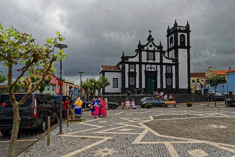 Square with historic church Nossa Senhora da Luz in the background, people in traditional dress, dark clouds and parked cars, Fenais da Luz, Sao Miguel Island, Azores, Portugal, Europe