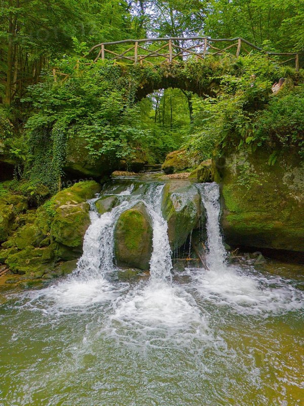 An idyllic waterfall flows under a rustic bridge, surrounded by lush greenery and rocks, aerial view, waterfall Schiessentümpel, Schéissendëmpel, Schwarze Ernz, Mühlental, Müllerthal, Mullerthal, Waldbillig, Luxembourg, Europe