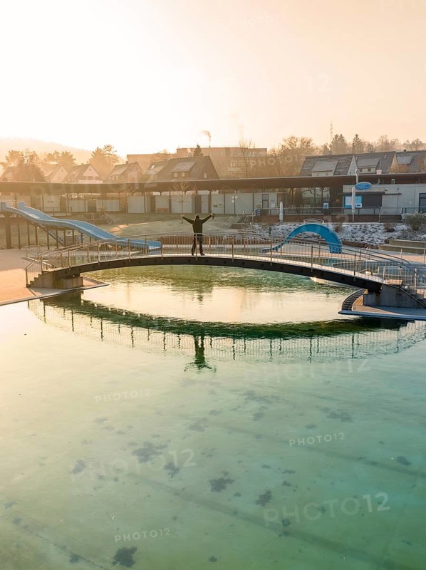A person stands on a bridge over a swimming pool with green frozen water in the winter sun, ENCW Freibad Calw, Stammheim, Black Forest, Germany, Europe