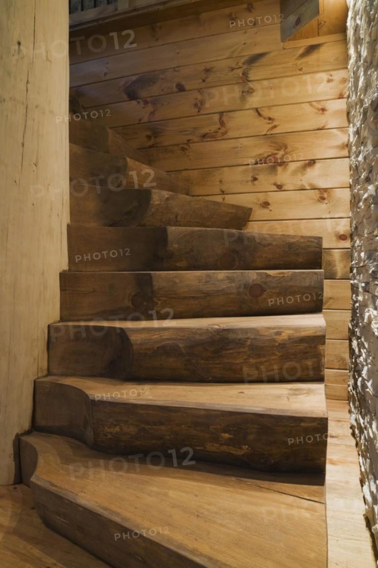 Illuminated twisting wooden staircase with brown stained half log steps attached to large bleached supporting central tree trunk post plus illuminated lights shining on tan natural stone and wood plank wall in basement leading to ground floor inside handcrafted Eastern white pine Scandinavian log cabin home, Quebec, Canada, North America