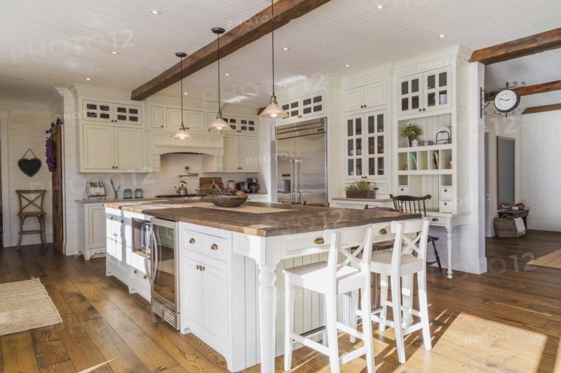 White painted maple wood antique style cabinets with tan coloured quartz countertops, island with brown stained wood plank countertop and high back bar stools in kitchen with stained oak wood floorboards inside country style home, Quebec, Canada, North America